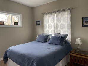 A bedroom with a bed, a nightstand, a lamp, and a window with curtains. - Spacious Frankfort Getaway - Walk to Downtown and Lake Michigan - Sleeps 12-14. (Frankfort)