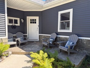 A house exterior with a porch, four Adirondack chairs, a white door, and a window. - Spacious Frankfort Getaway - Walk to Downtown and Lake Michigan - Sleeps 12-14. (Frankfort)