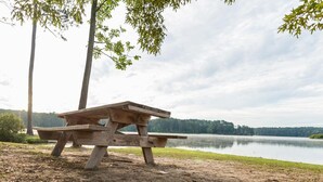 A wooden picnic table by a lake with trees in the background. - Studio 6 Meridian, MS - Bonita Lakes (Meridian)