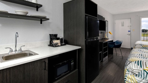 A compact kitchenette with a sink, microwave, and black refrigerator. There are shelves above the sink holding white cups and bowls. A small desk with a chair is next to the refrigerator. - Studio 6 Meridian, MS - Bonita Lakes (Meridian)