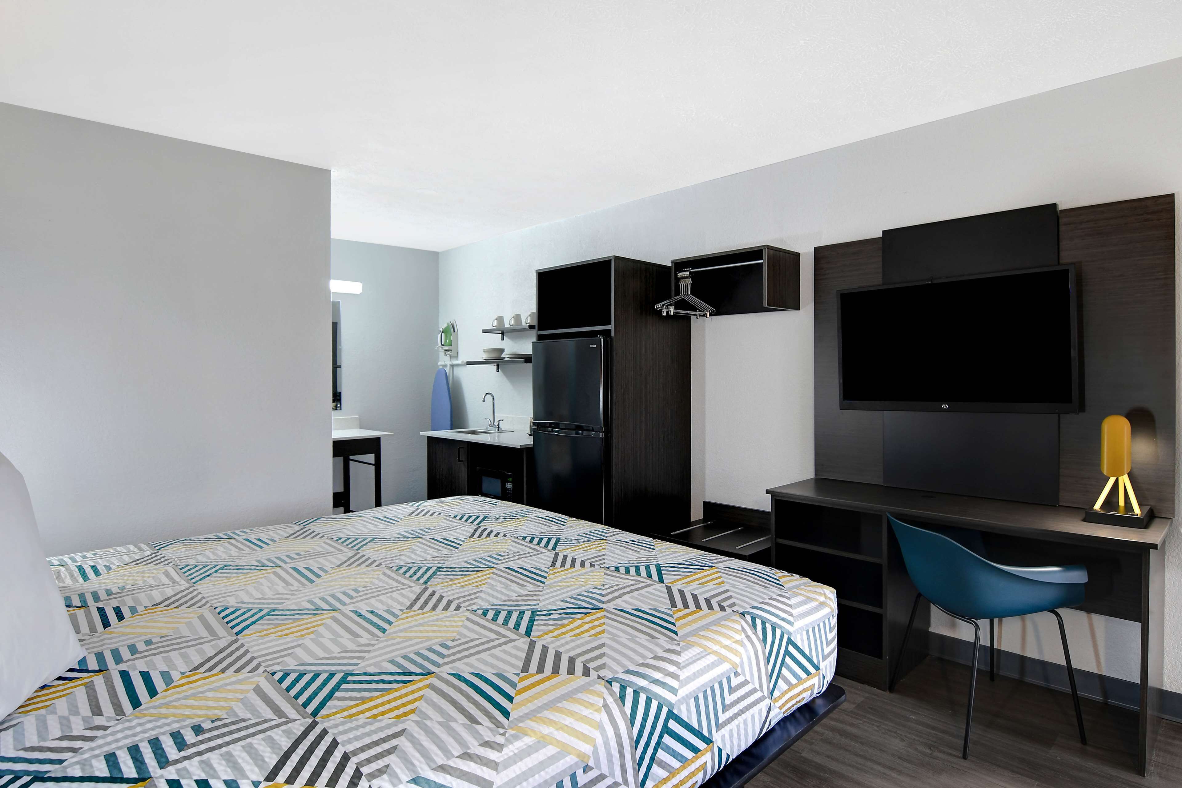 Quarto de hotel moderno com cama, escrivaninha, cadeira, TV e cozinha compacta.