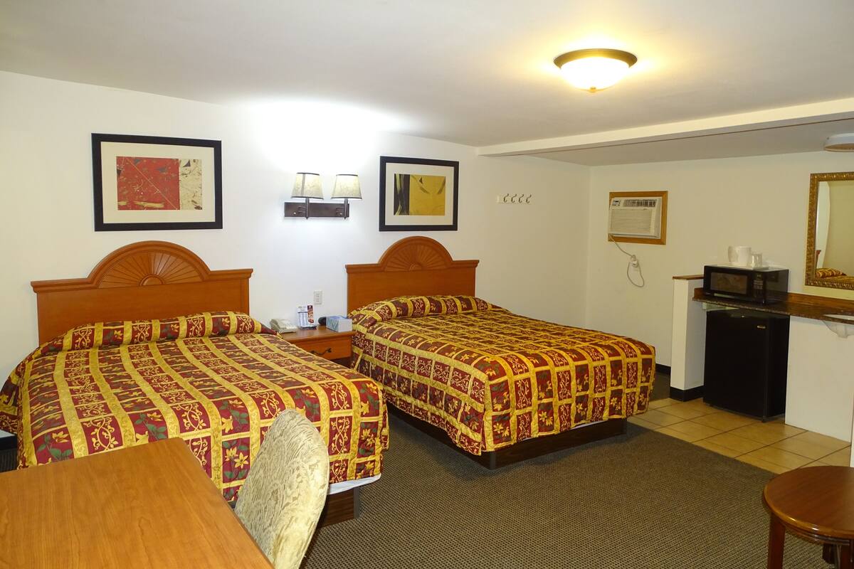 A hotel room with two beds, a desk, a chair, a microwave, an air conditioner, and framed artwork on the walls.