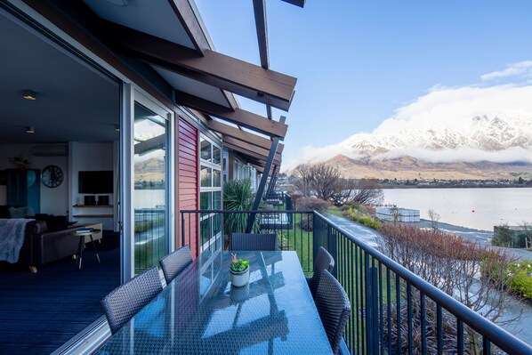Uma varanda com mesa e cadeiras de vidro, com vista para um lago e montanhas.