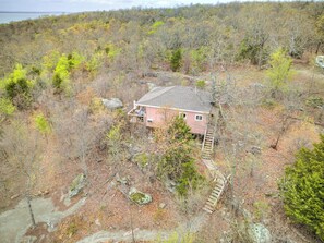 A single-story house with a sloped roof, surrounded by a forested area with a dirt path leading to the entrance.