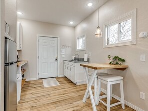 A compact kitchen with a wooden floor, white cabinetry, a small dining area with a wooden table and white stool, and a window with a view of bare trees.