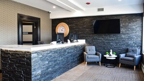 A reception area with a black brick wall, a large round clock, a flat-screen TV, and two grey armchairs with a small table between them. - Big Bird Inn Wawa, SureStay Collection by Best Western (Wawa)
