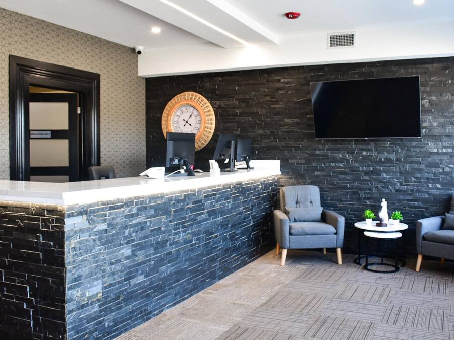 Un espace d’accueil avec un mur en briques noires, une grande horloge ronde, un téléviseur à écran plat et deux fauteuils gris disposés de part et d’autre d’une petite table.