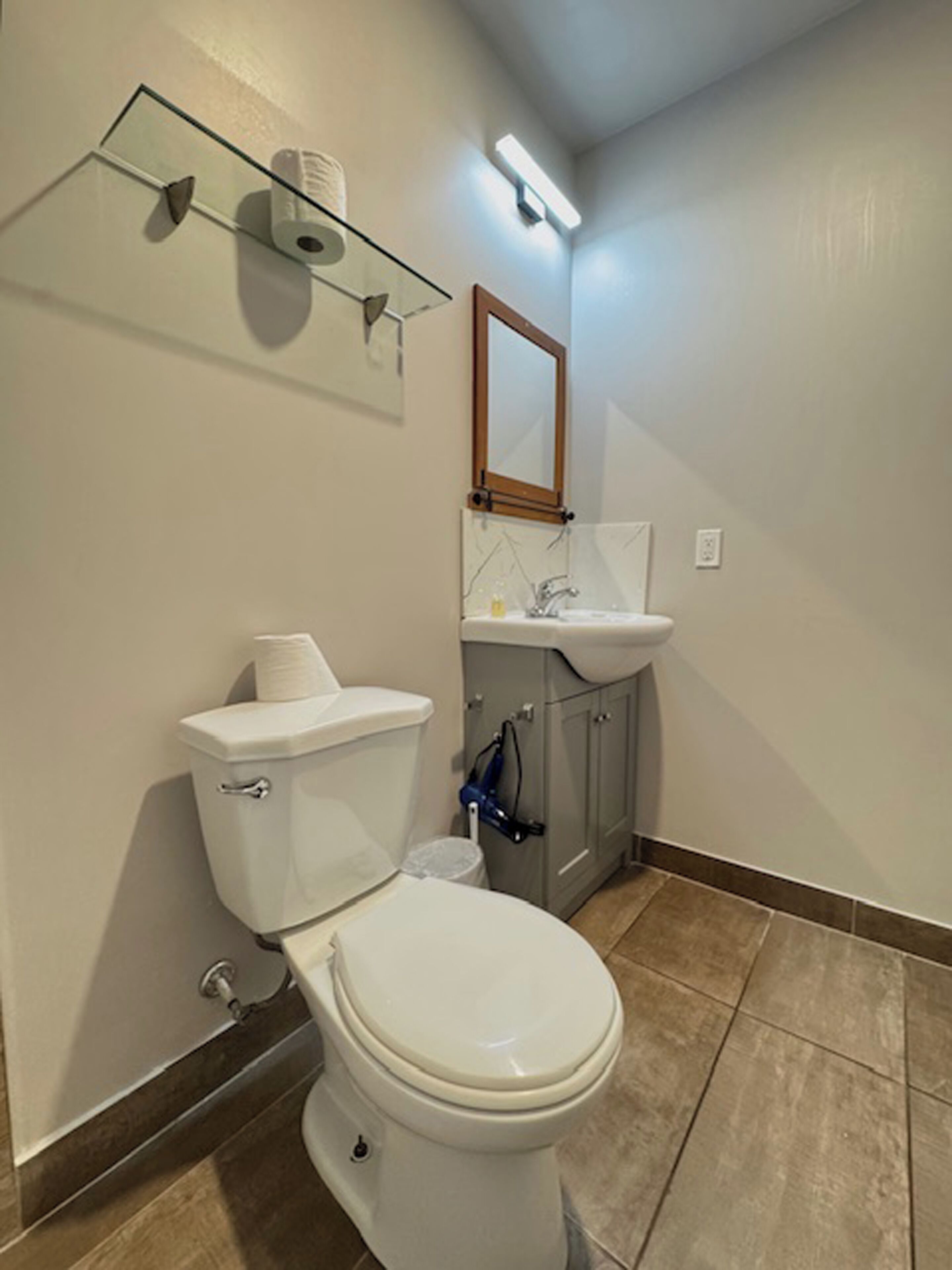 Un baño con inodoro blanco, estante de vidrio con papel higiénico, espejo sobre el lavabo y suelo de baldosas.
