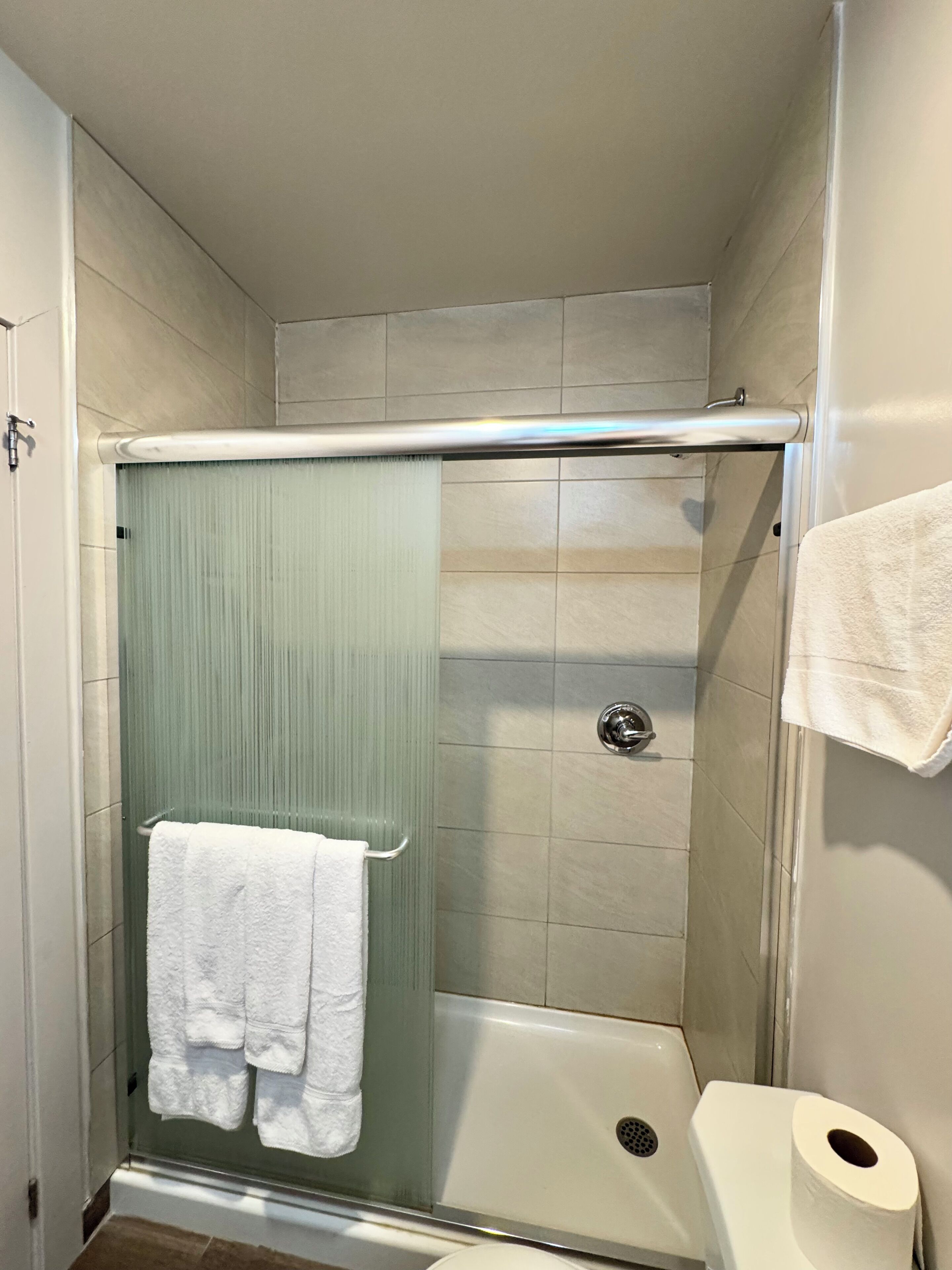 Une cabine de douche avec une porte en verre, une serviette blanche accrochée à la porte et des toilettes dotées d’un distributeur d’essuie-mains en papier.