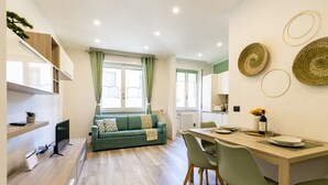 A modern living space with a sofa, dining table, and chairs. There are shelves with plants and a small TV. - Green Pearl ☆☆☆☆☆ - Apartment 100 meters from the lake - WiFi - By Guestaly (Arona)