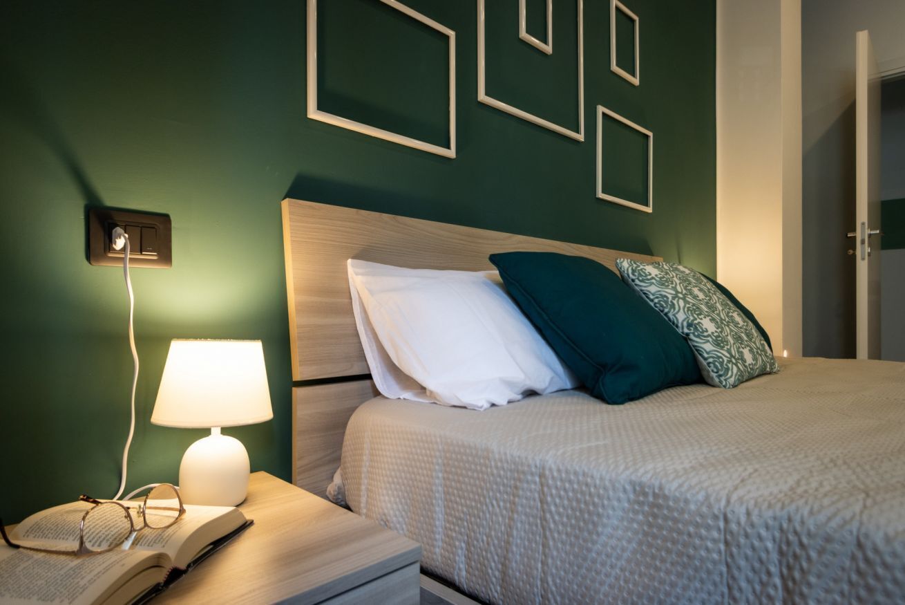 Un lit bien fait, avec un oreiller blanc et turquoise, une lampe de chevet et un livre ouvert sur une table de chevet.