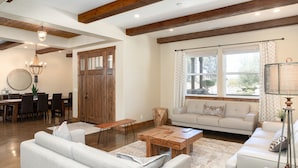 A spacious living room with a wooden ceiling, a dining area with a round table and chairs, and a large window with curtains.