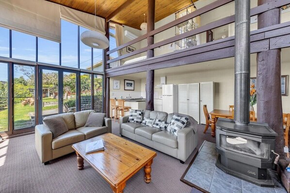 A spacious living area with a high ceiling, a large window, a sofa, a coffee table, and a wood-burning stove. - Studio Views for Two People (Mallacoota)