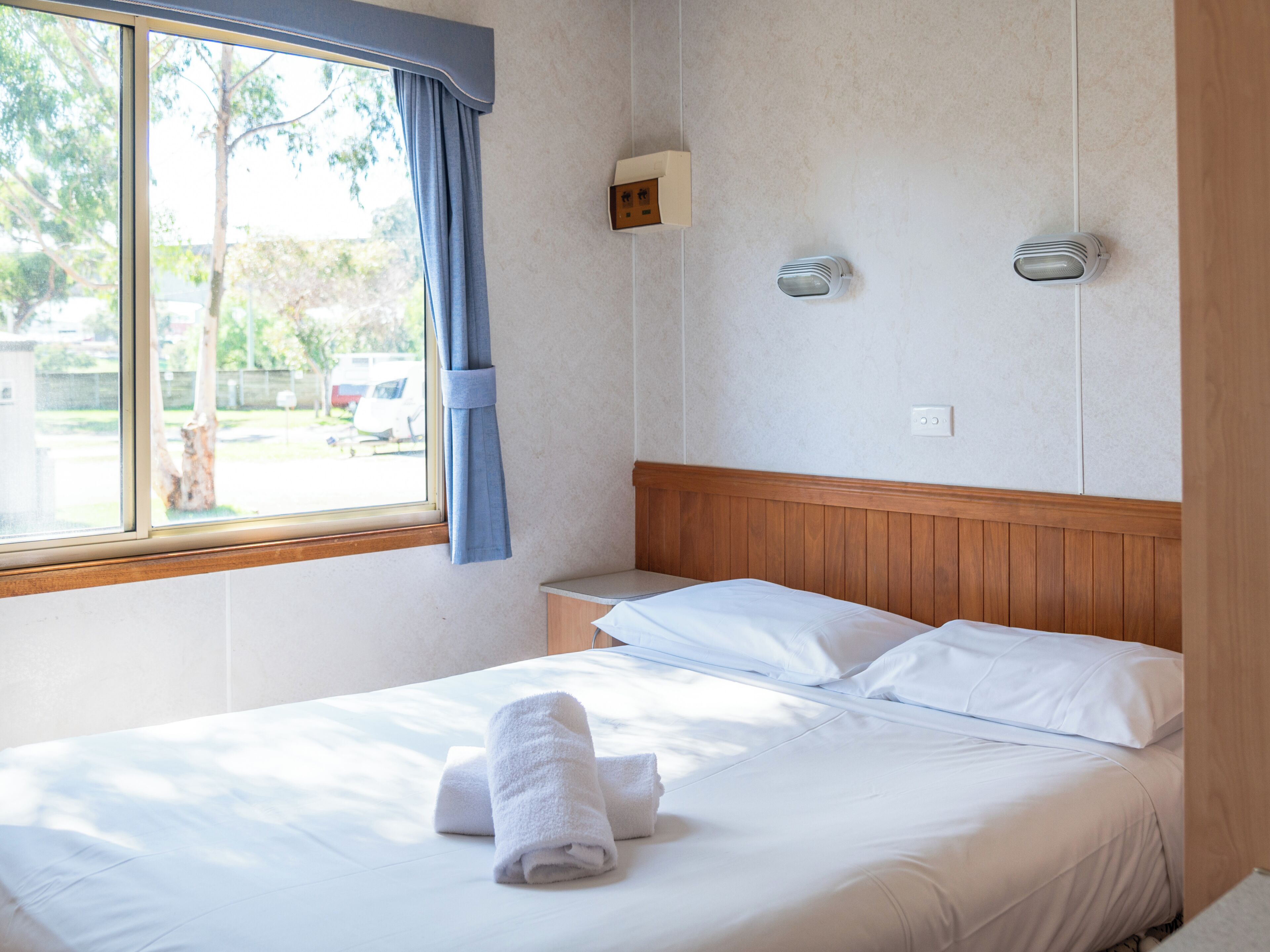 A hotel room with a bed, a nightstand, a window with curtains, and a wall-mounted light fixture.
