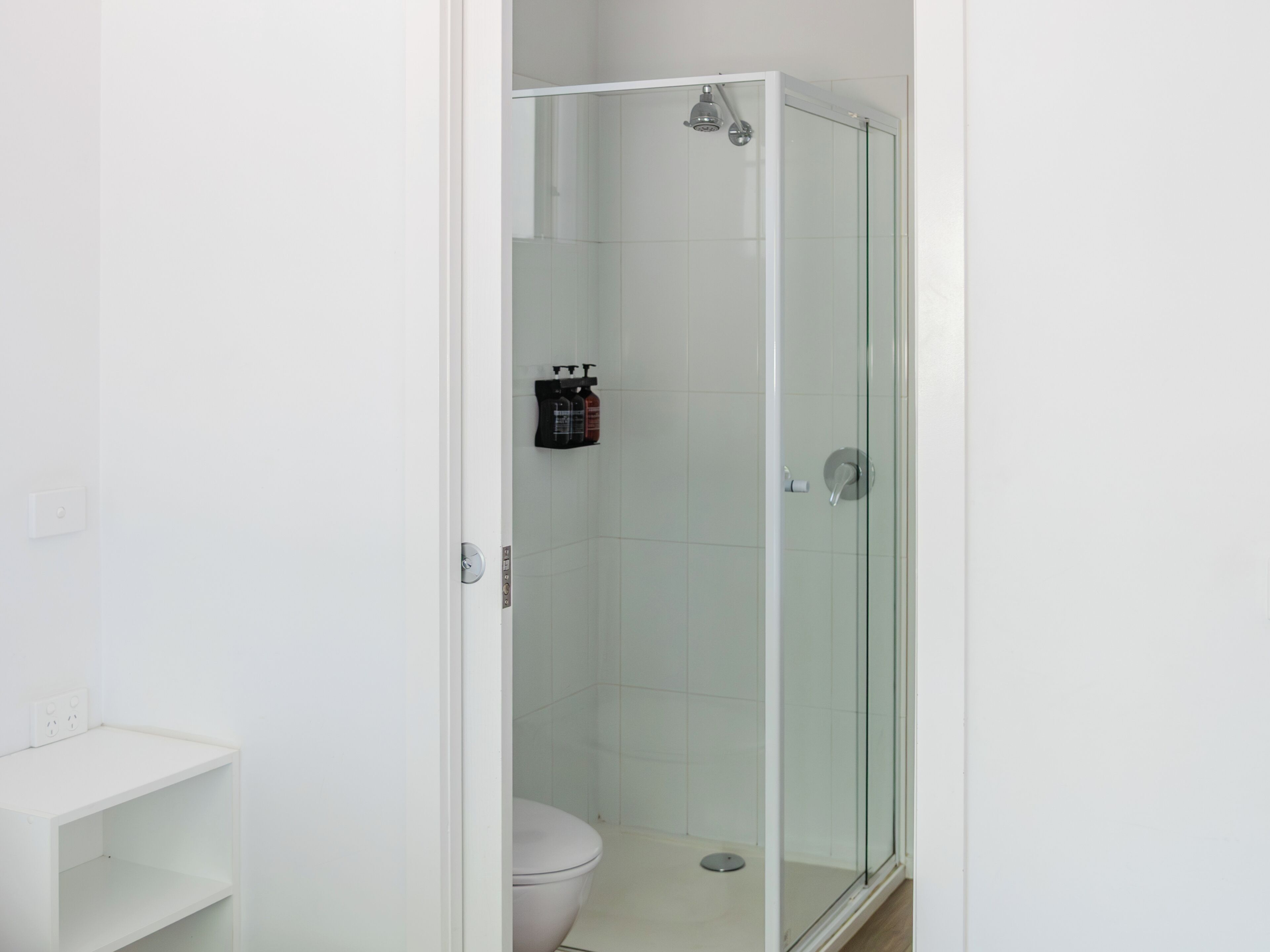 A bathroom with a glass-enclosed shower, a white toilet, and a shelf with toiletries.