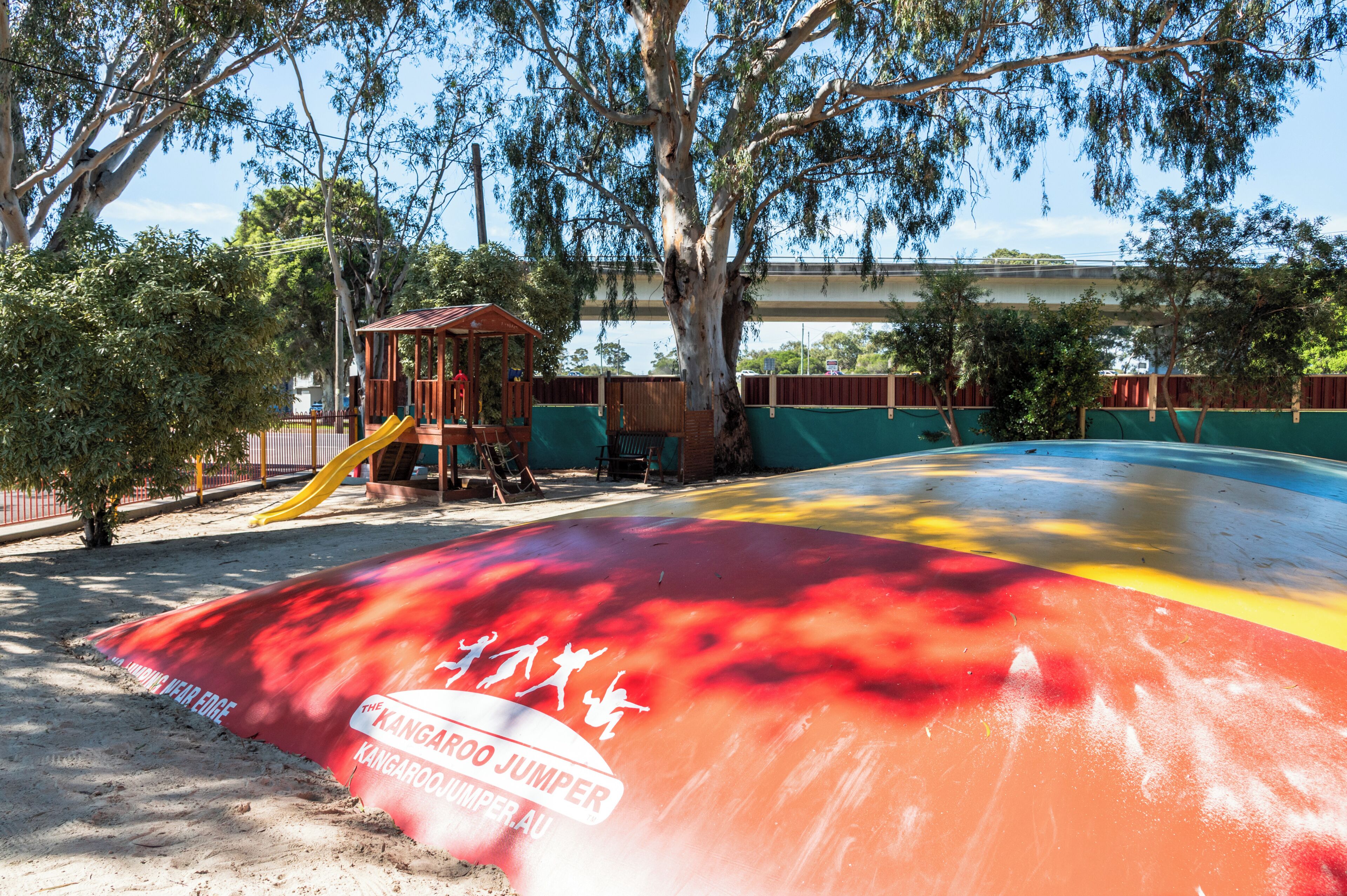 Un parque infantil con tobogán y estructuras para escalar, rodeado de árboles y cercado.
