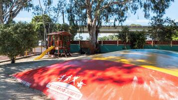 Un parque infantil con tobogán y estructuras para escalar, rodeado de árboles y cercado.