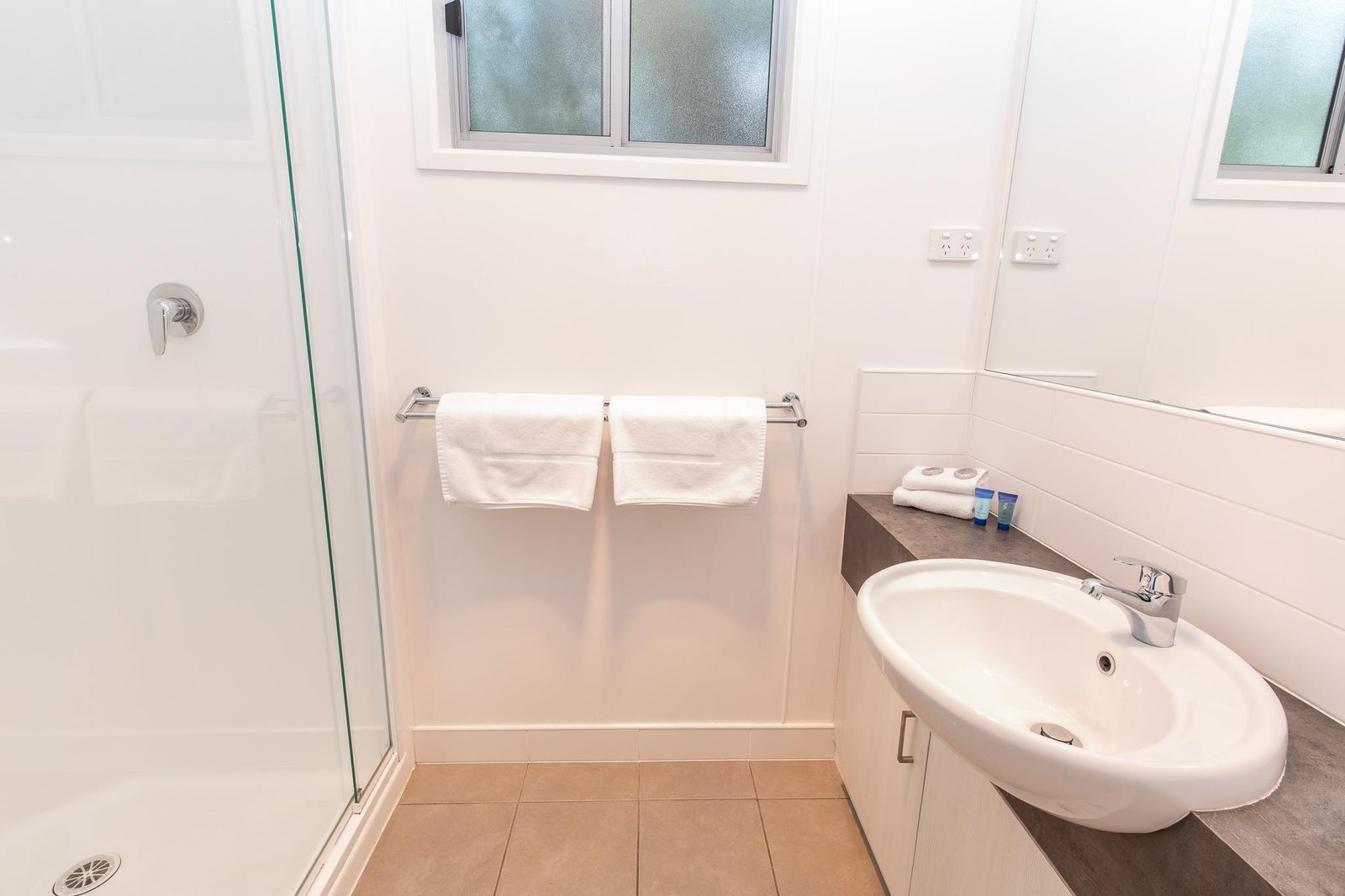Un baño moderno con cabina de ducha de vidrio, un lavabo blanco y una ventana sobre el espejo.