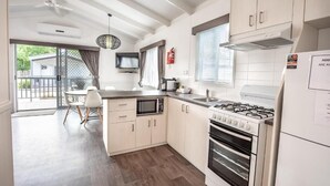 A modern kitchen with a gas stove, microwave, and white cabinets. There is a dining area with a table and chairs, and a large window with blinds. - Geelong Holiday Park (Belmont)