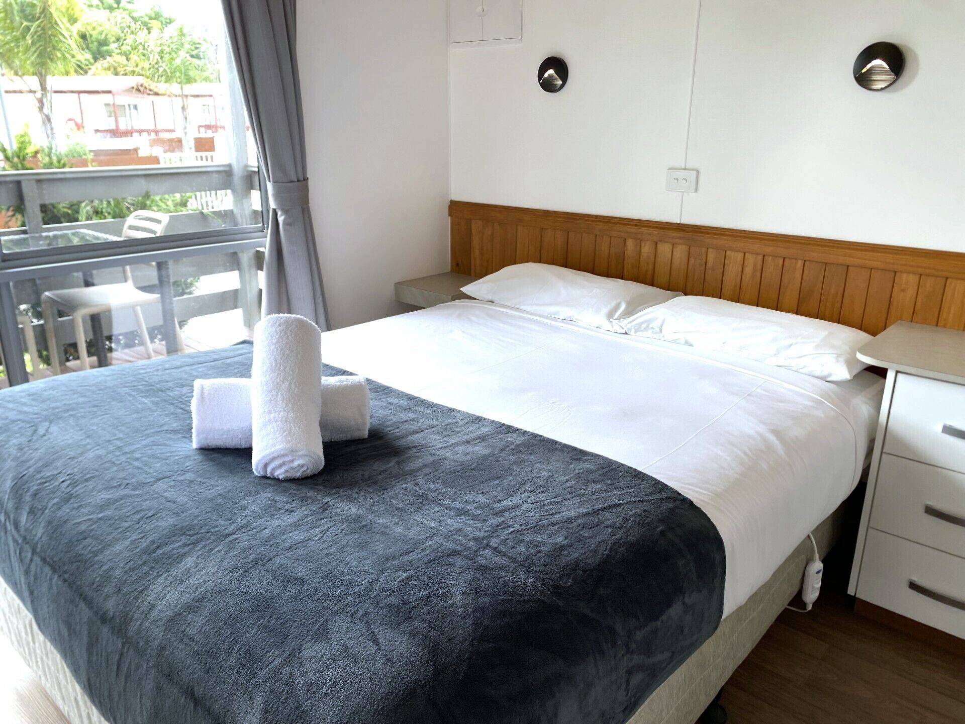 Un letto rifatto con cura, una coperta grigia e due asciugamani arrotolati, una testiera in legno, un comodino con una lampada, e vista all'esterno attraverso una finestra.