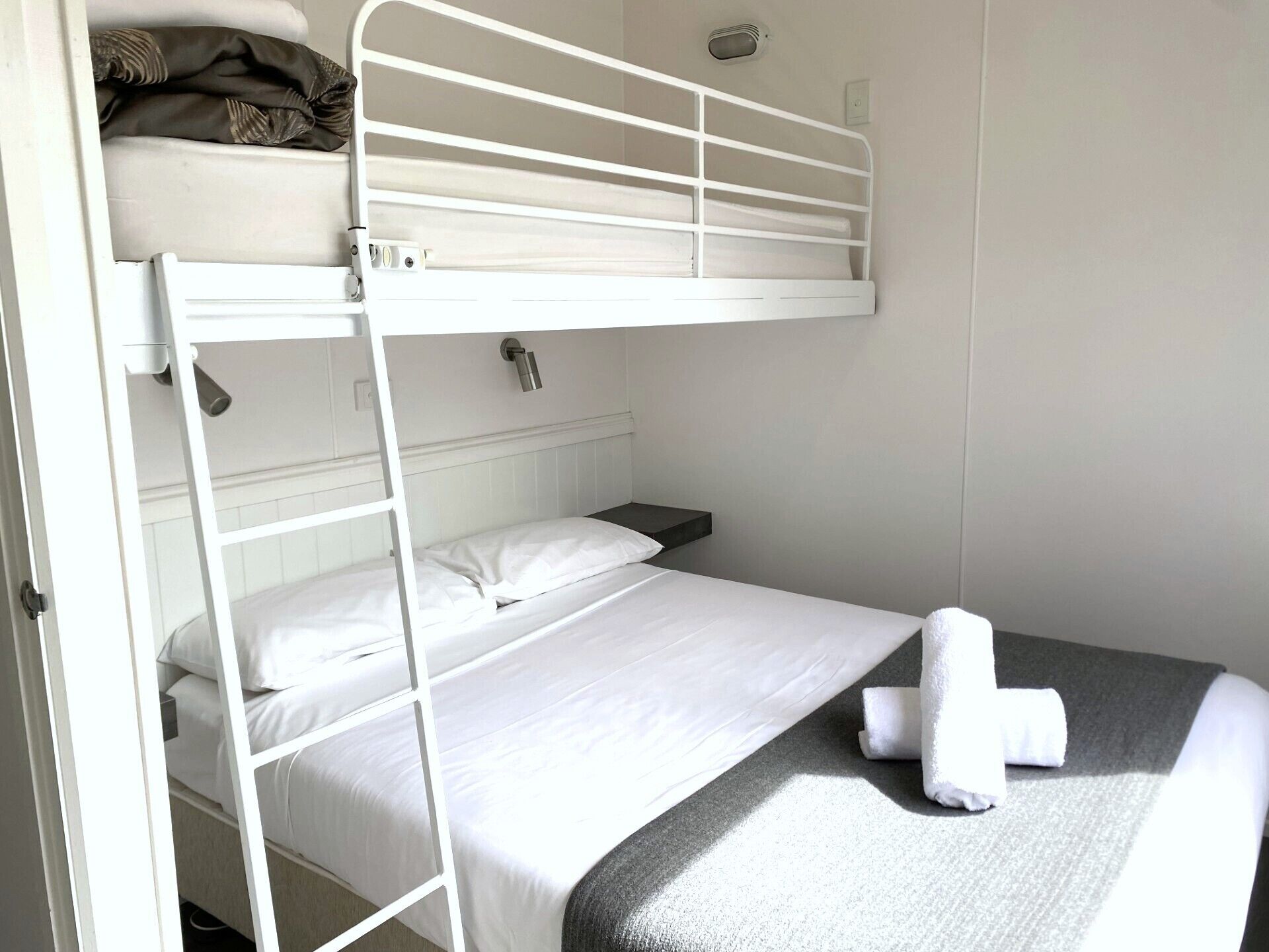 Habitación con literas, cabecera blanca, mesita de noche y una toalla enrollada en la litera inferior.