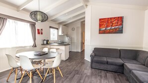 A modern living room with a grey sofa, a dining table with white chairs, and a kitchen area in the background. - Geelong Holiday Park (Belmont)