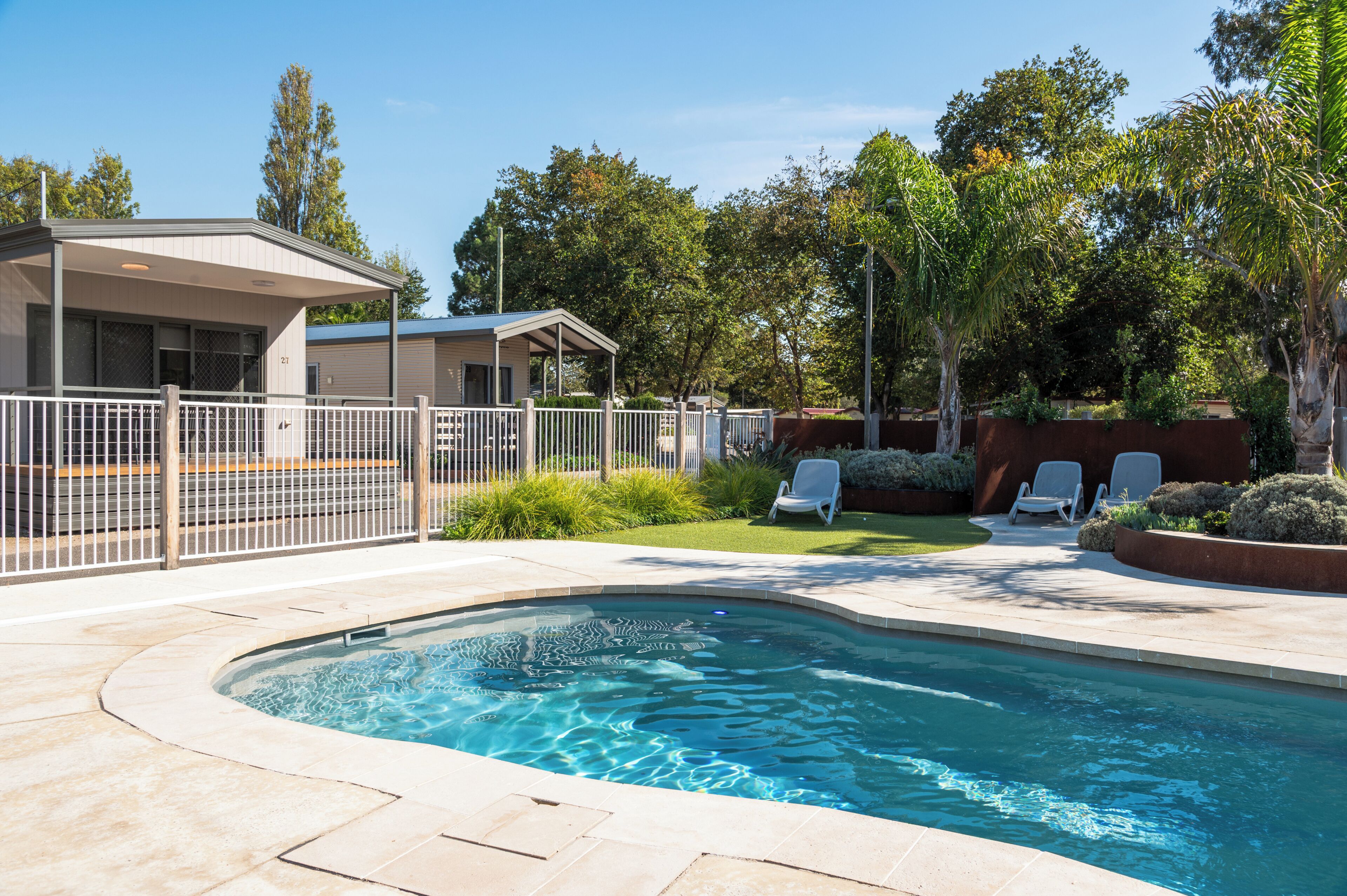 A swimming pool with a tiled deck, a fenced area with lounge chairs, and a building with a covered patio.