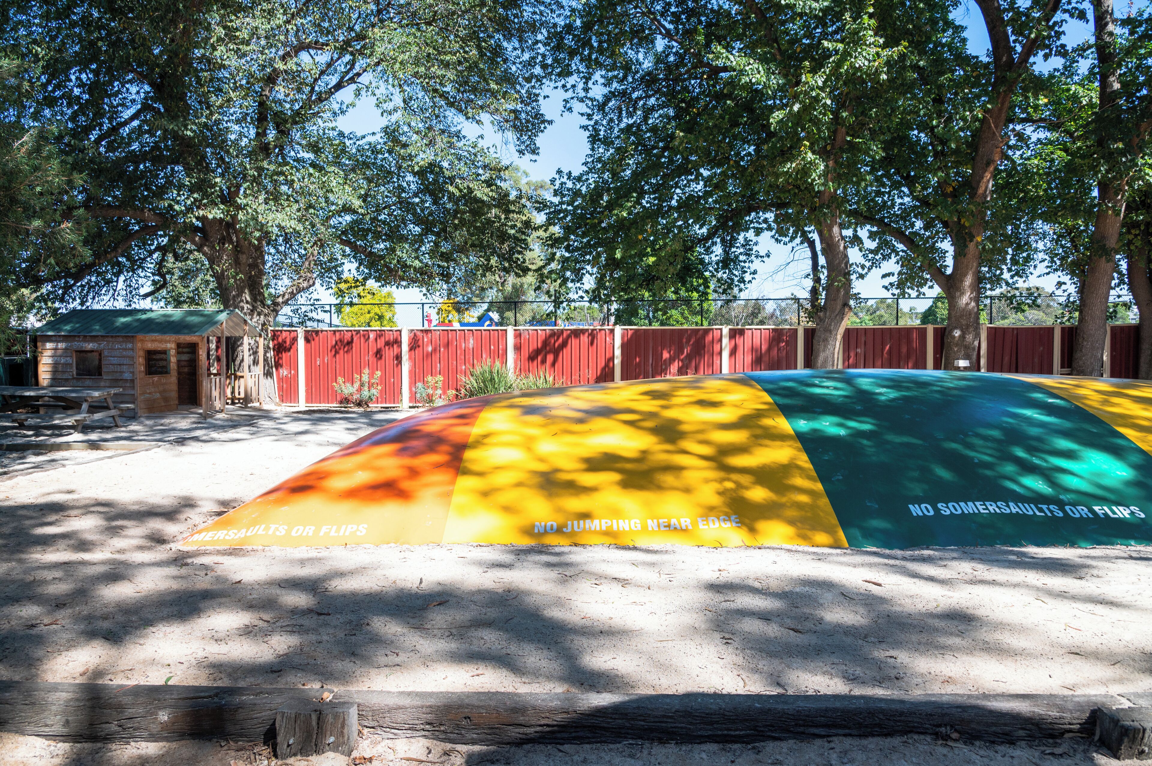 A colorful trampoline with safety warnings, surrounded by trees and a wooden fence.