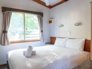 A hotel room with a large bed, a window with curtains, a bedside table, and a wall-mounted light fixture. - Geelong Holiday Park (Belmont)
