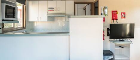 A compact kitchen with white cabinets, a microwave, and a stove. There's a TV on a stand and a trash bin.