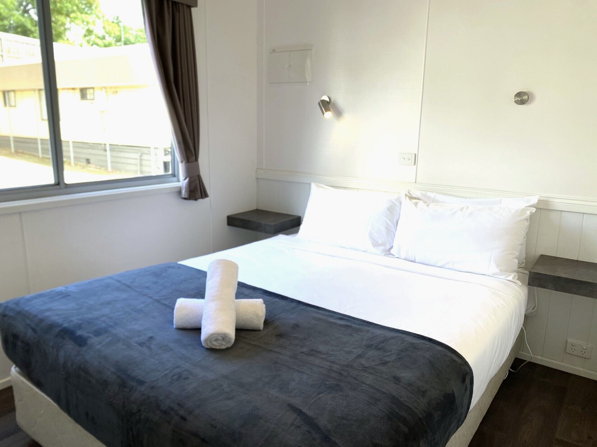 A hotel room with a bed, a bedside table, a window with curtains, and a view of the outside.