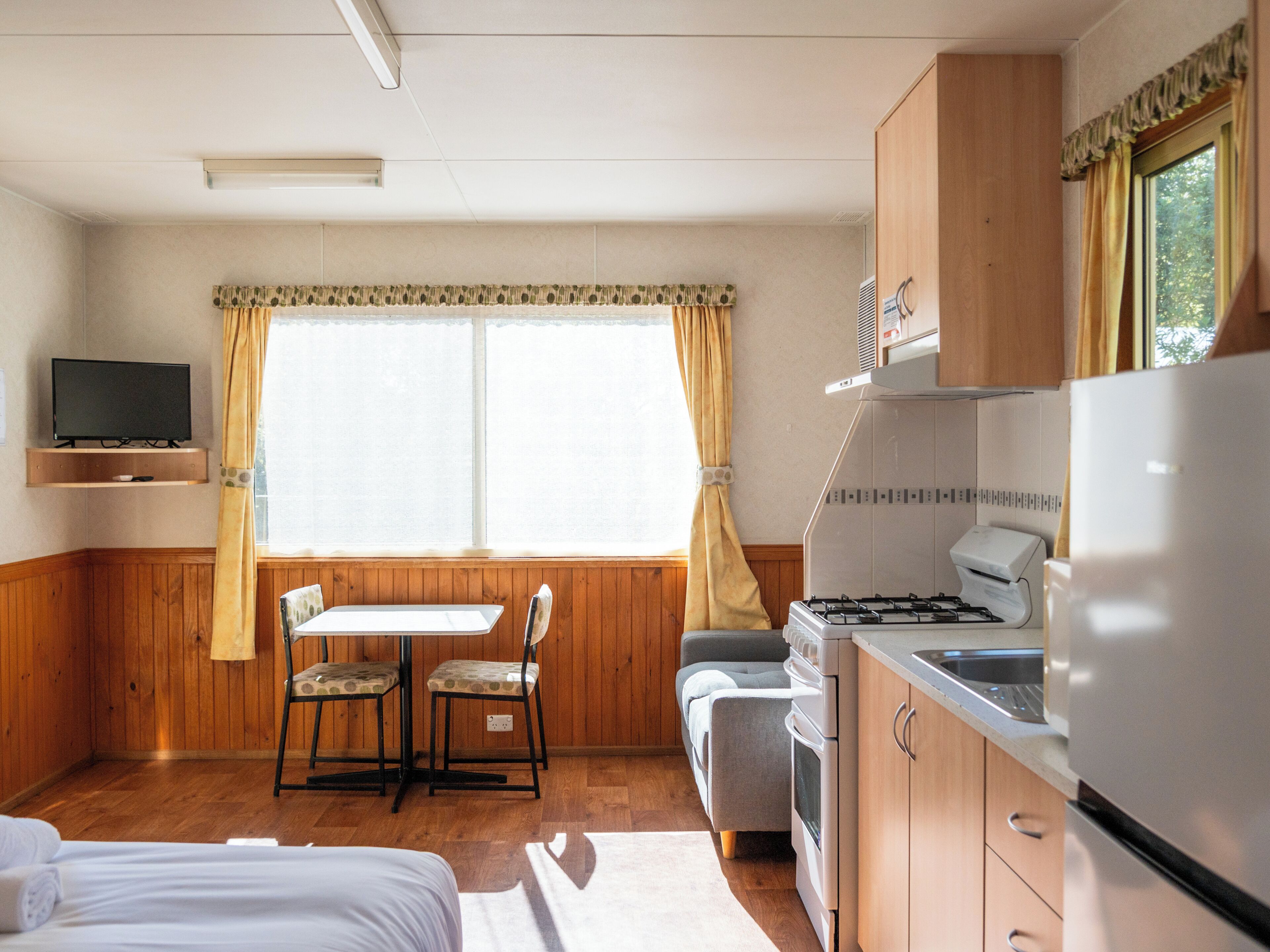 A compact room with a bed, a small table with chairs, a kitchenette with a stove and refrigerator, and a television on the wall.