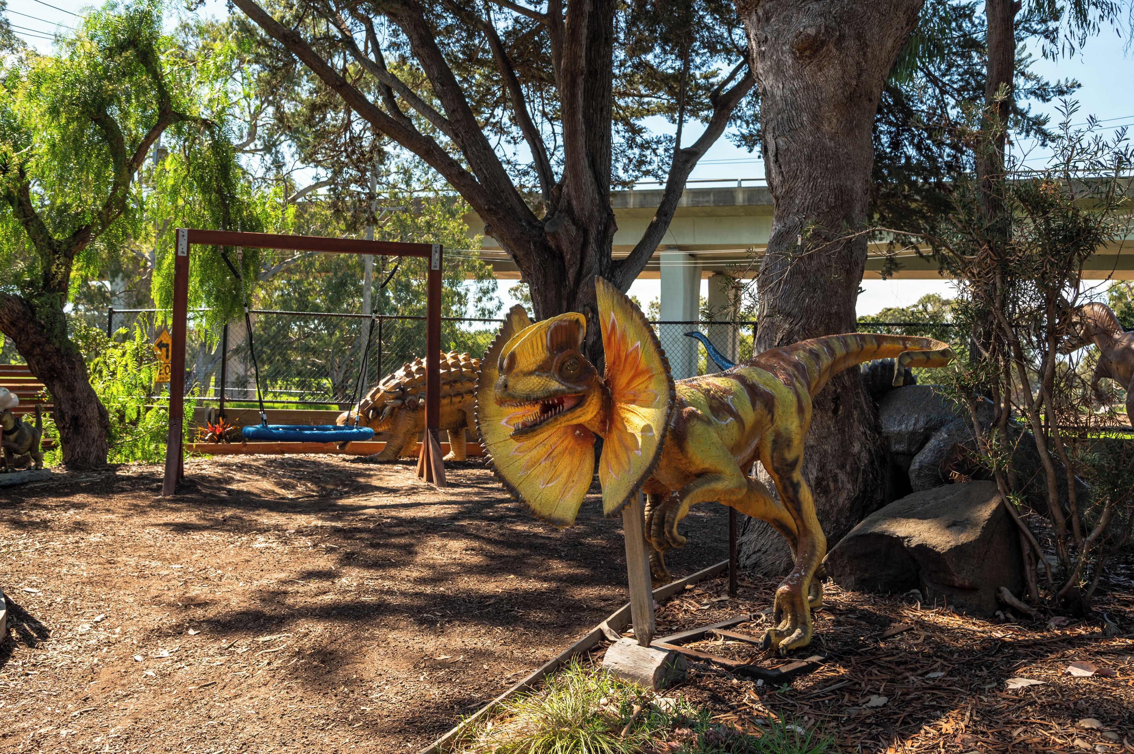 Een dinosaurus tentoonstelling met een groot dinosaurusmodel op de voorgrond, omringd door bomen en een schommel in de verte.