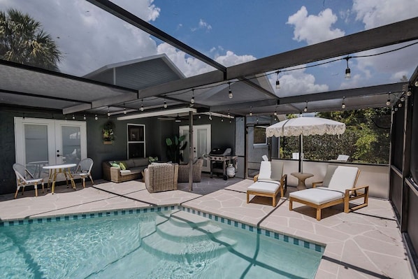 A covered outdoor area with a pool, lounge chairs, and a grill.