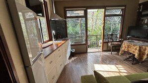A spacious living area with a wooden floor, a green sofa, a dining table with a patterned tablecloth, a kitchen area with a refrigerator, and a sliding glass door leading to a balcony with a view of trees. - Blue Mountains studio, wild native garden full of birds, week-long stay 30% off (Hazelbrook)