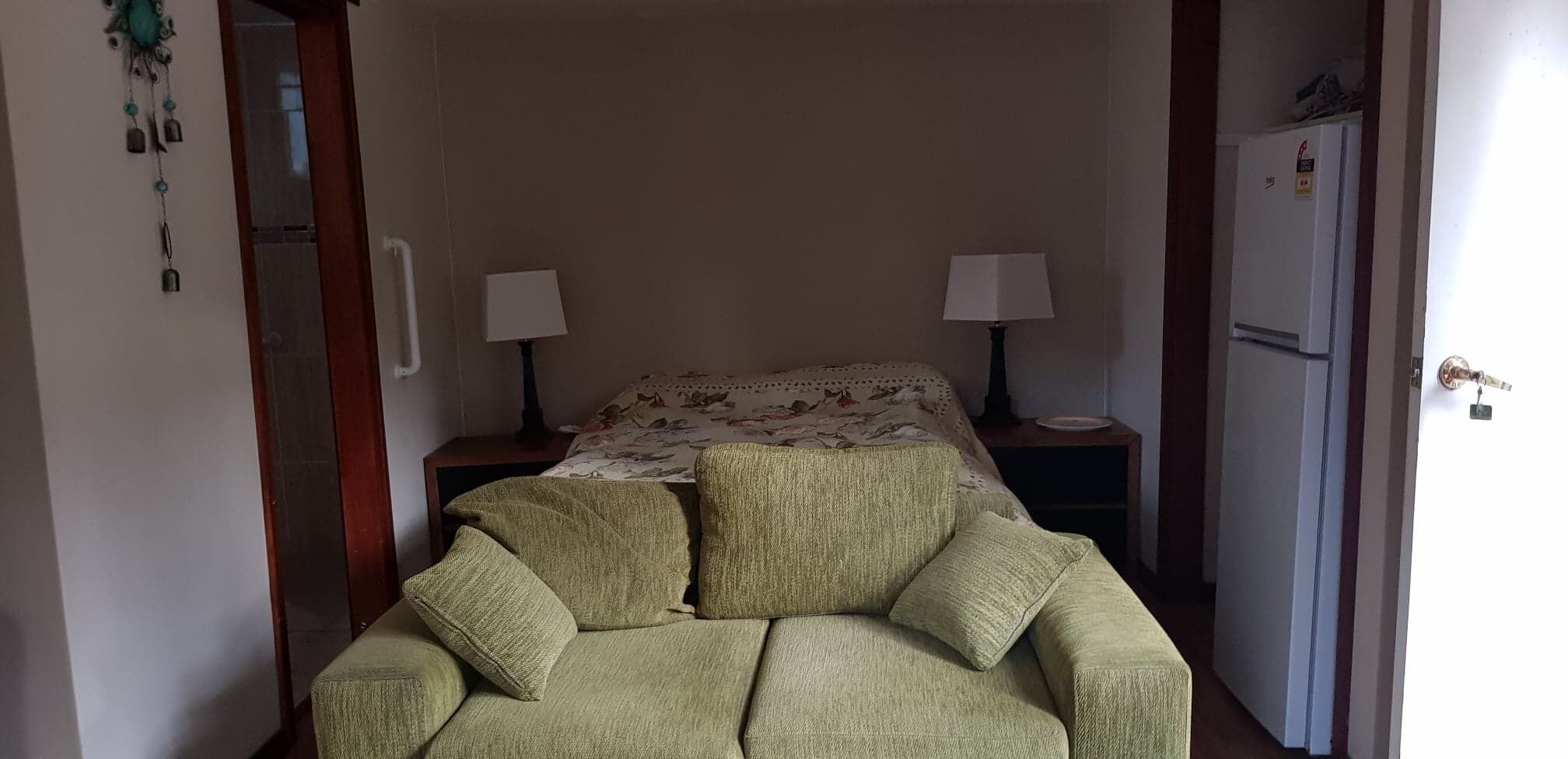 A small, cozy room with a green sofa, two bedside tables with lamps, a bed with floral bedding, and a refrigerator.