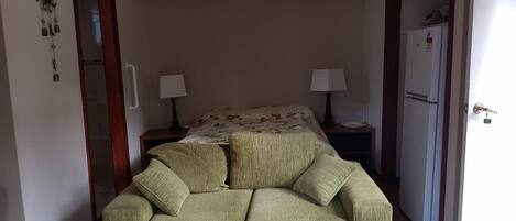 A small, cozy room with a green sofa, two bedside tables with lamps, a bed with floral bedding, and a refrigerator.