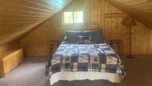 Um quarto de sótão acolhedor, com teto inclinado, uma cama com edredom xadrez, uma mesinha de cabeceira, um abajur e uma janela que deixa entrar luz natural.