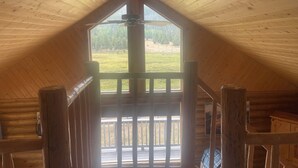 Uma cabana de madeira com teto inclinado, um ventilador de teto central e vista para um campo gramado através de uma janela grande.
