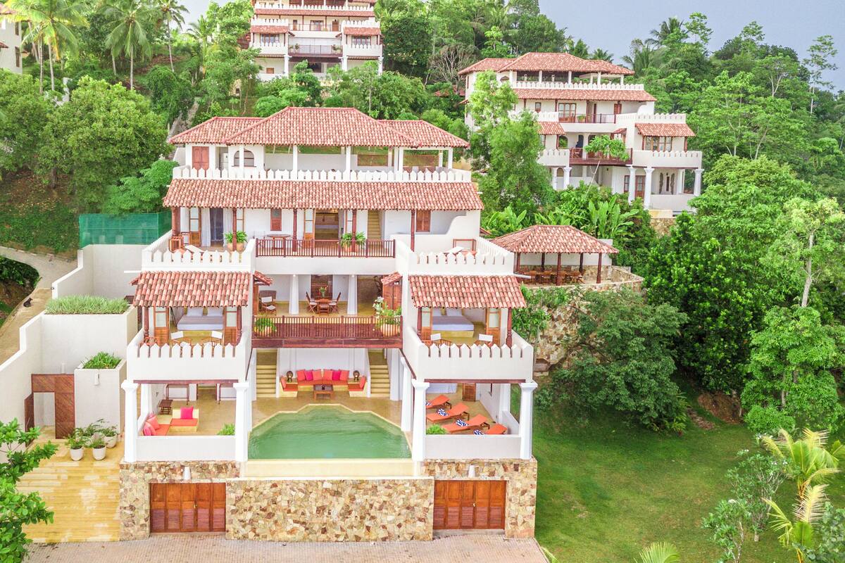 Un hotel de varios pisos con un patio central, balcones y abundante vegetación que rodea la propiedad.