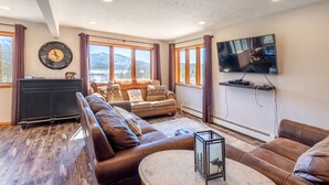 Una sala de estar con un sofá, una mesa de centro, un televisor montado en la pared y vistas a montañas a través de amplios ventanales.