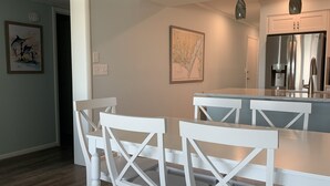 A modern kitchen with white cabinetry, a stainless steel refrigerator, and a dining area with white chairs and a marble countertop. - Shore Beats Work! Direct Oceanfront Atlantic Beach Paradise (Atlantic Beach)