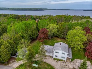 Une maison blanche avec un toit gris, entourée d’arbres verdoyants et touffus, et une étendue d’eau en arrière-plan.