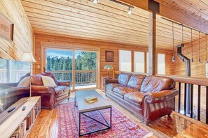 A spacious living room with wooden ceilings and large windows, a leather sofa, a coffee table, and a bookshelf.