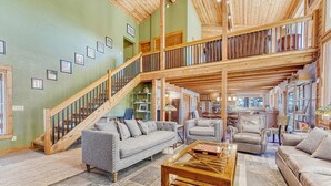 Ein großzügiger Wohnbereich mit hoher Decke, einer Holztreppe und verschiedenen Sitzgelegenheiten, darunter Sofas und Sessel.