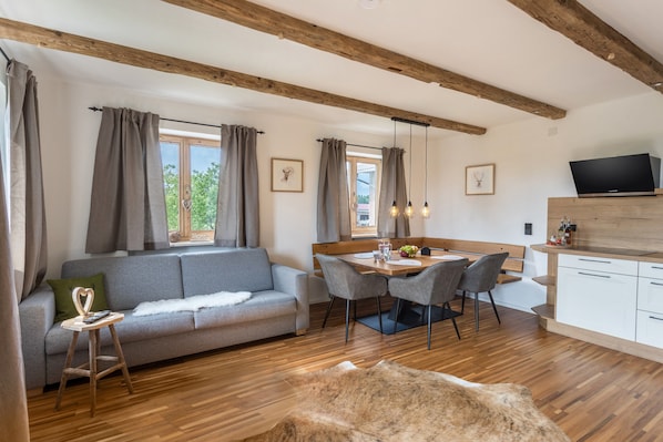 A modern living room with a sofa, dining table, chairs, wooden beams, and a TV.