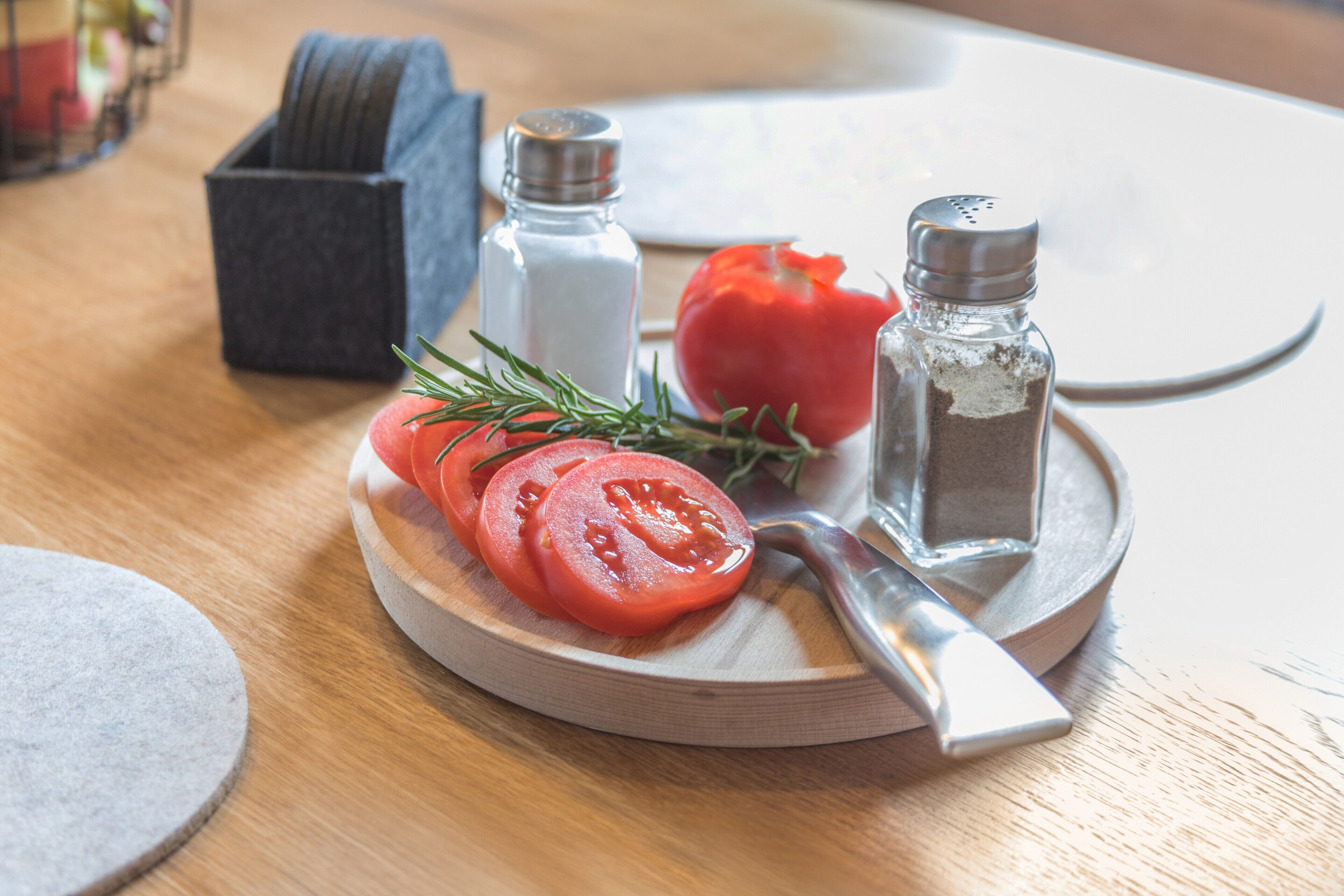 Une planche à découper en bois sur laquelle on voit des tomates coupées en tranches, un couteau, une branche de romarin et deux salières, le tout posé sur une table en bois.