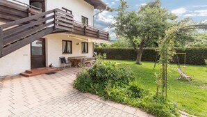 Ein zweistöckiges Haus mit einer Holztreppe, einer Terrasse mit Tisch und Stühlen sowie einem gepflegten Garten mit Schaukel.