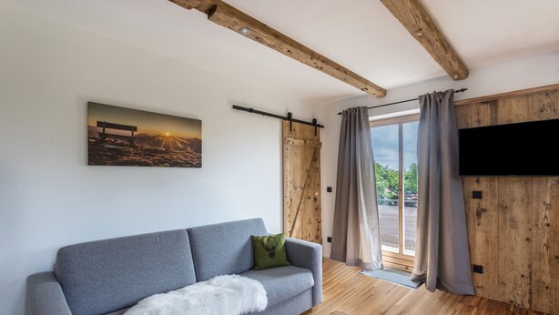 Ein modernes Wohnzimmer mit grauem Sofa, einer Wand-Fernseher, Holzbalken, einem großen Fenster mit Vorhängen und einem Bild an der Wand.