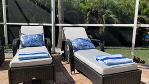 Zwei Korbsessel mit weißen Kissen und blau gemusterten Polstern, auf einer gefliesten Terrasse mit Blick auf Palmen und einem klaren Himmel.